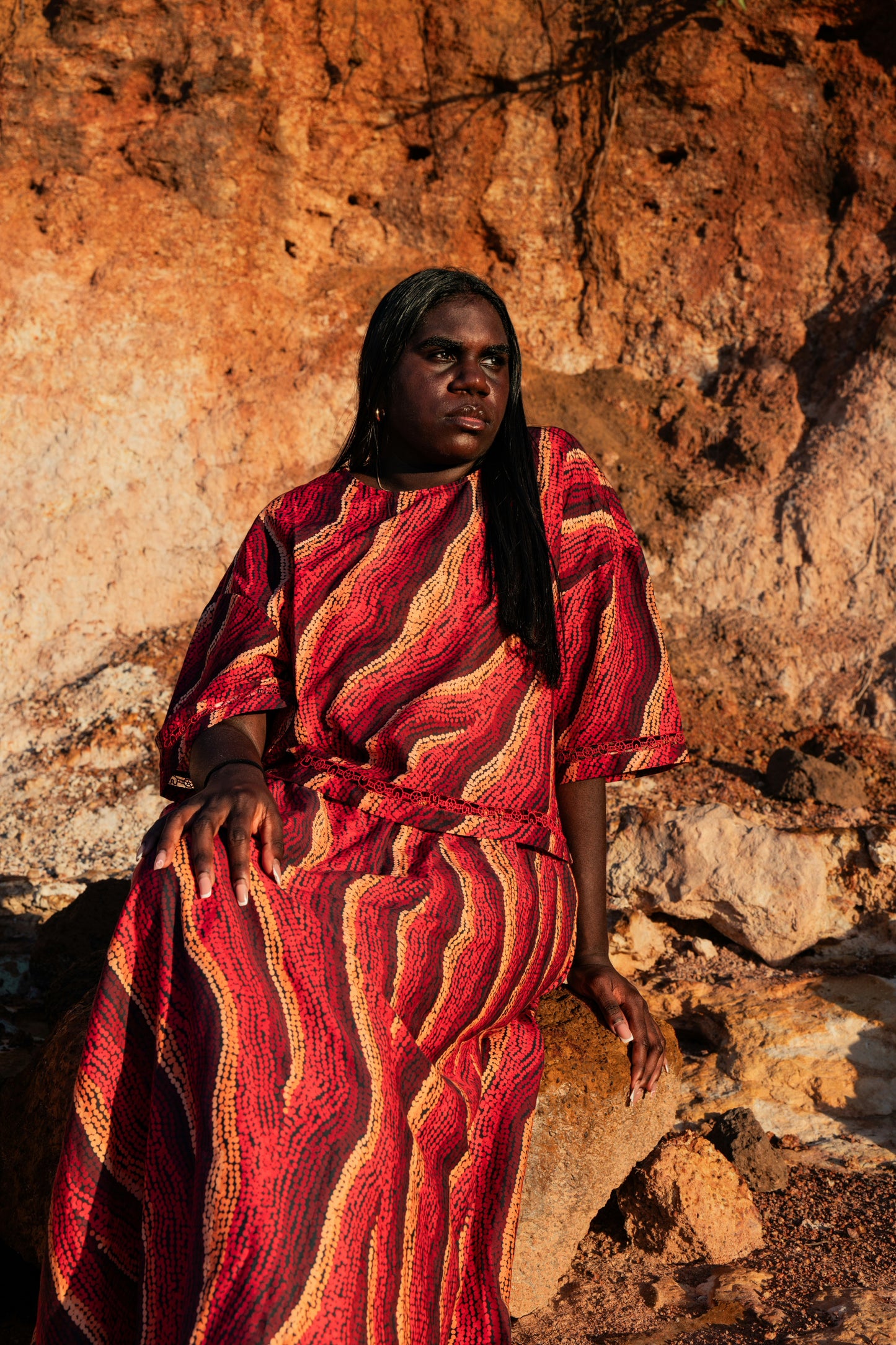 An Aboriginal woman  wearing a tshirt and top with Aboriginal artwork against a rocky background