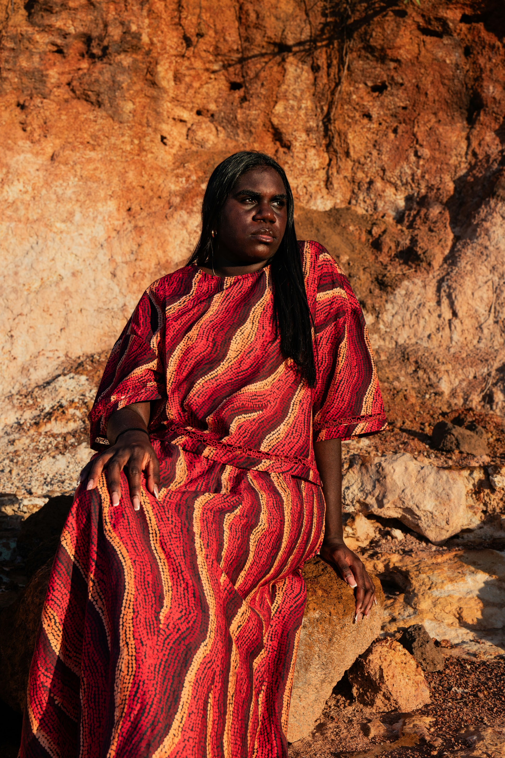 An Aboriginal woman  wearing a tshirt and top with Aboriginal artwork against a rocky background
