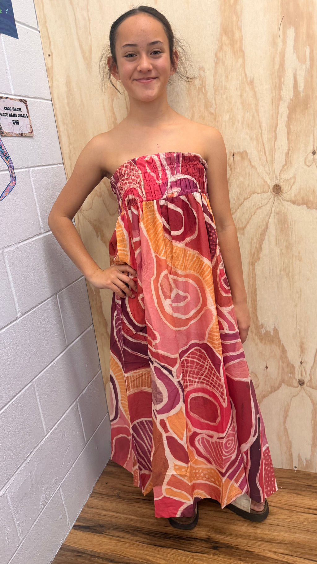 A young girl stands in a corner wearing a strapless maxi dress with a vibrant Aboriginal pattern in shades of pink, red, orange, and purple. The background consists of a white brick wall on the left, a wooden panel wall on the right, and a wooden floor.