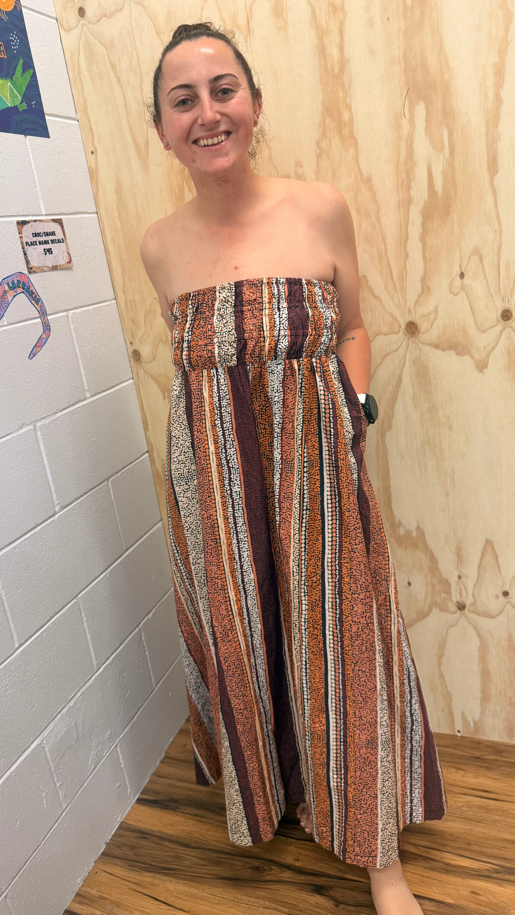 A woman wearing a strapless maxi dress with vertical Aboriginal art pattern brown, rust, beige, and white stripes, standing in a room with a white brick wall, wood paneling, and a wood floor.