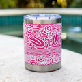 A stainless steel insulated tumbler with a bright pink and white Aboriginal patterned wrap, featuring a clear lid with blue trim, set on a stone ledge with a blurred swimming pool and green foliage in the background.