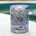 A stainless steel tumbler with a black and white Aboriginal pattern, featuring a clear lid with a blue tint, sitting on a stone surface with a blurred pool and greenery background.