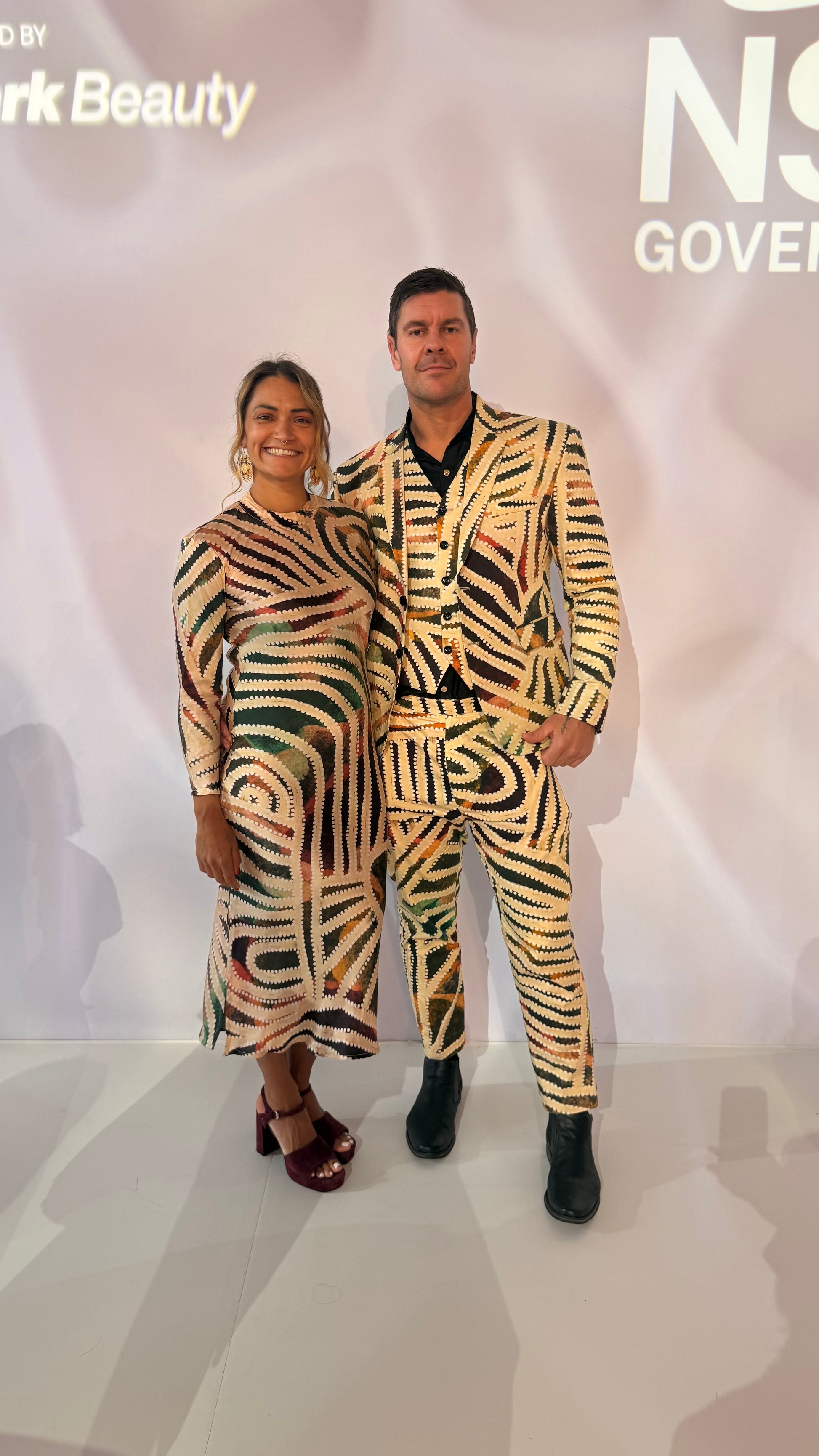 Two people in matching Aboriginal artwork outfits standing in front of a AFW sign