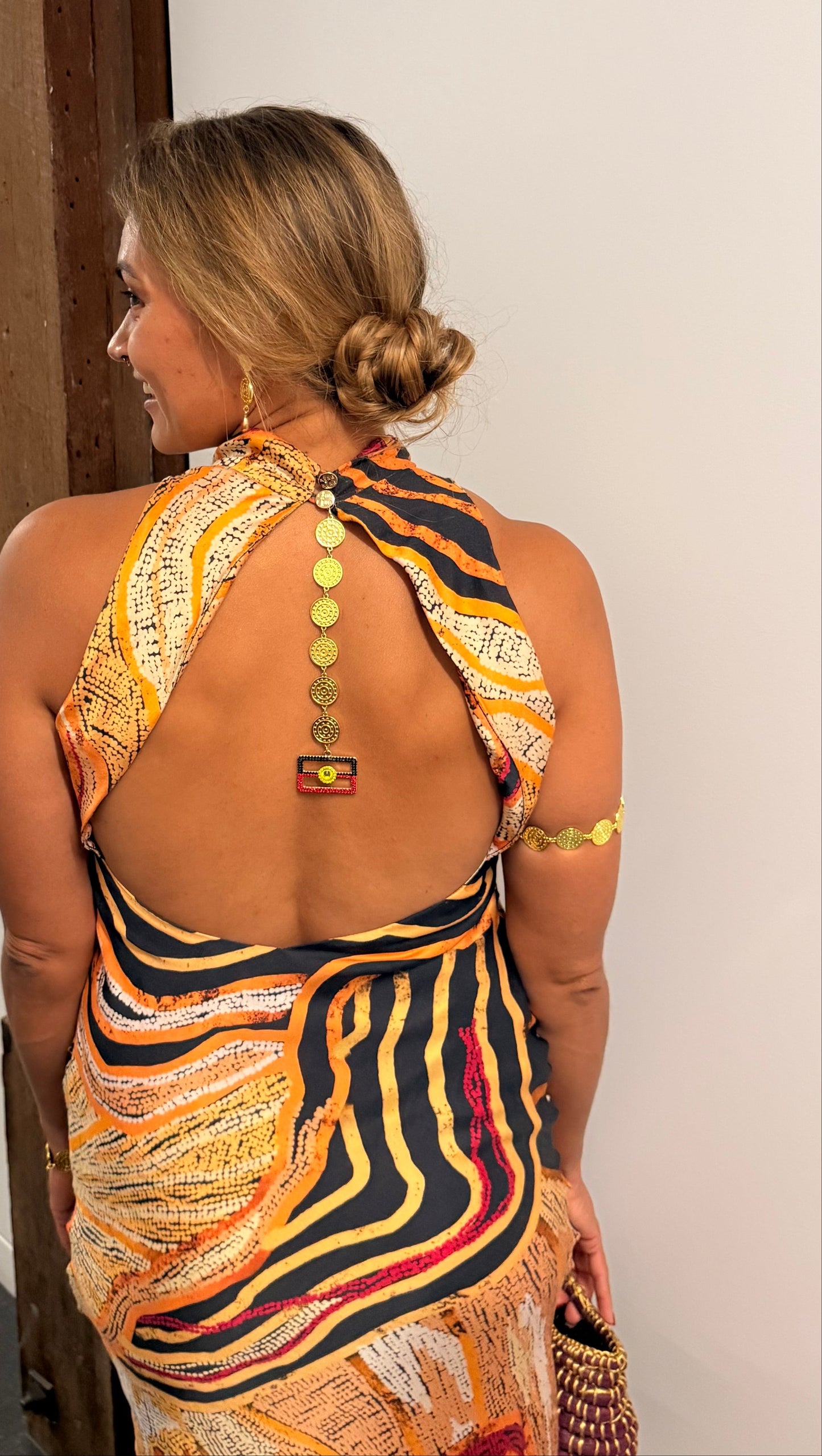 Aboriginal Woman in a First Nations designed dress with a open back and Aboriginal flag jewellery  standing against a white wall.