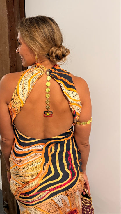 Aboriginal Woman in a First Nations designed dress with a open back and Aboriginal flag jewellery  standing against a white wall.
