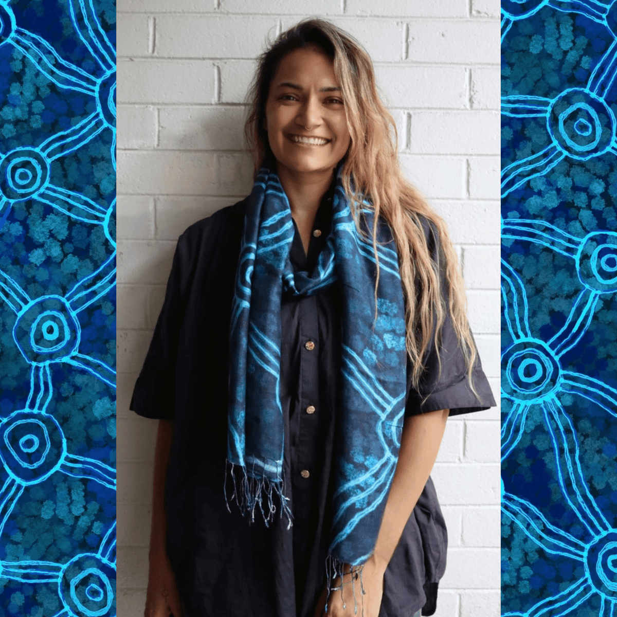 A Aboriginal woman wearing a brightly coloured blue scarf with Aboriginal artwork on it standing against a white brick wall