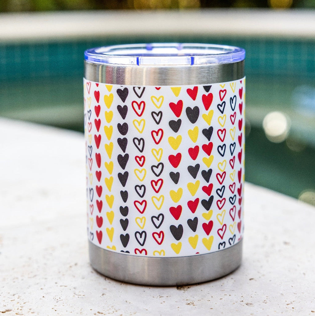 A stainless steel insulated tumbler with a clear lid sits on a light stone surface outdoors near a swimming pool. The tumbler is wrapped in a white sleeve covered with a repeating pattern of small hearts in red, yellow, black, and gray.