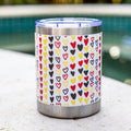 A stainless steel insulated tumbler with a clear lid sits on a light stone surface outdoors near a swimming pool. The tumbler is wrapped in a white sleeve covered with a repeating pattern of small hearts in red, yellow, black, and gray.