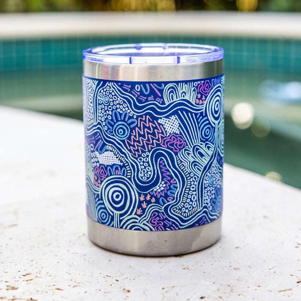 A stainless steel can cooler tumbler with a blue and purple Aboriginal pattern, sitting on a concrete ledge next to a swimming pool with a blurred green background.