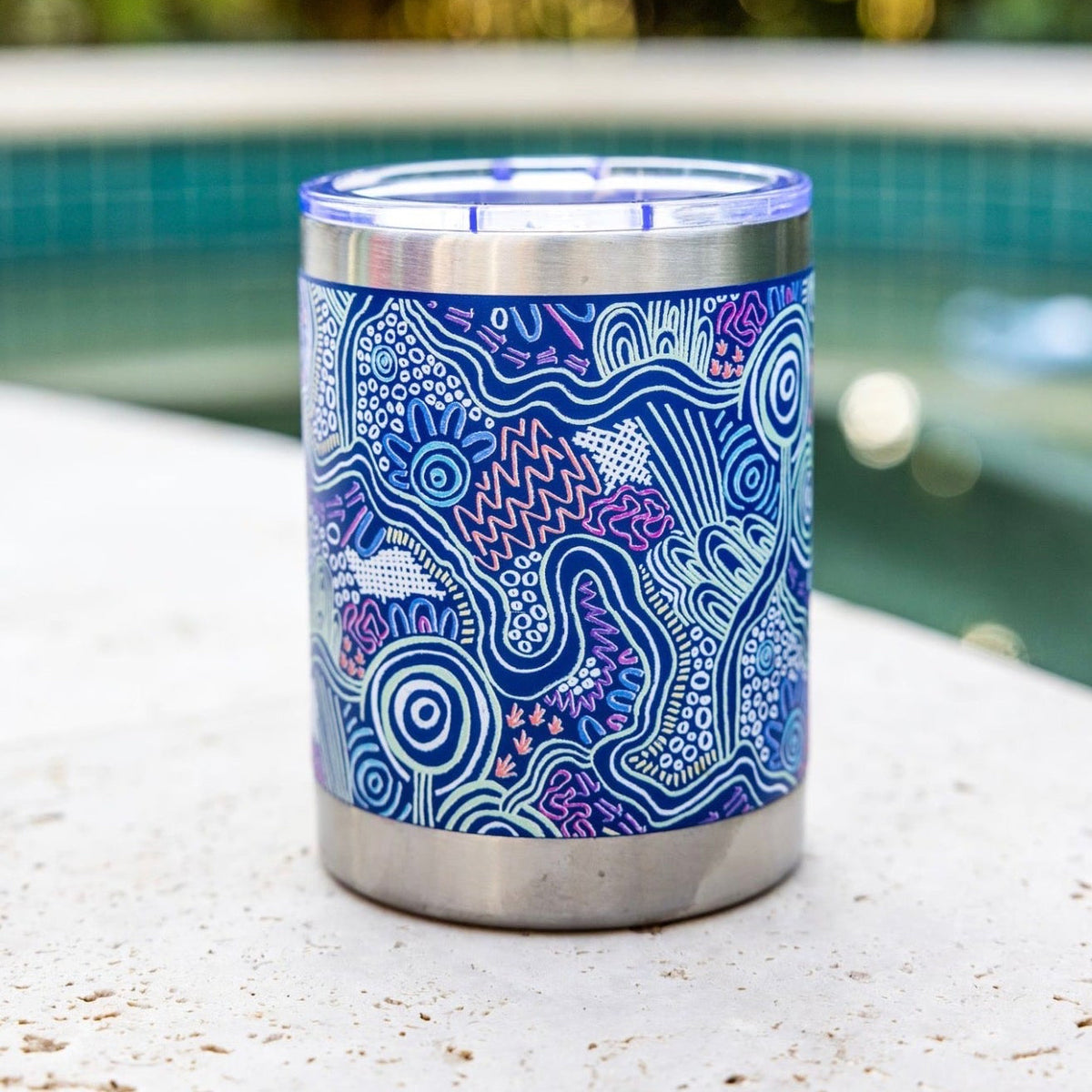 A stainless steel can cooler tumbler with a blue and purple Aboriginal pattern, sitting on a concrete ledge next to a swimming pool with a blurred green background.