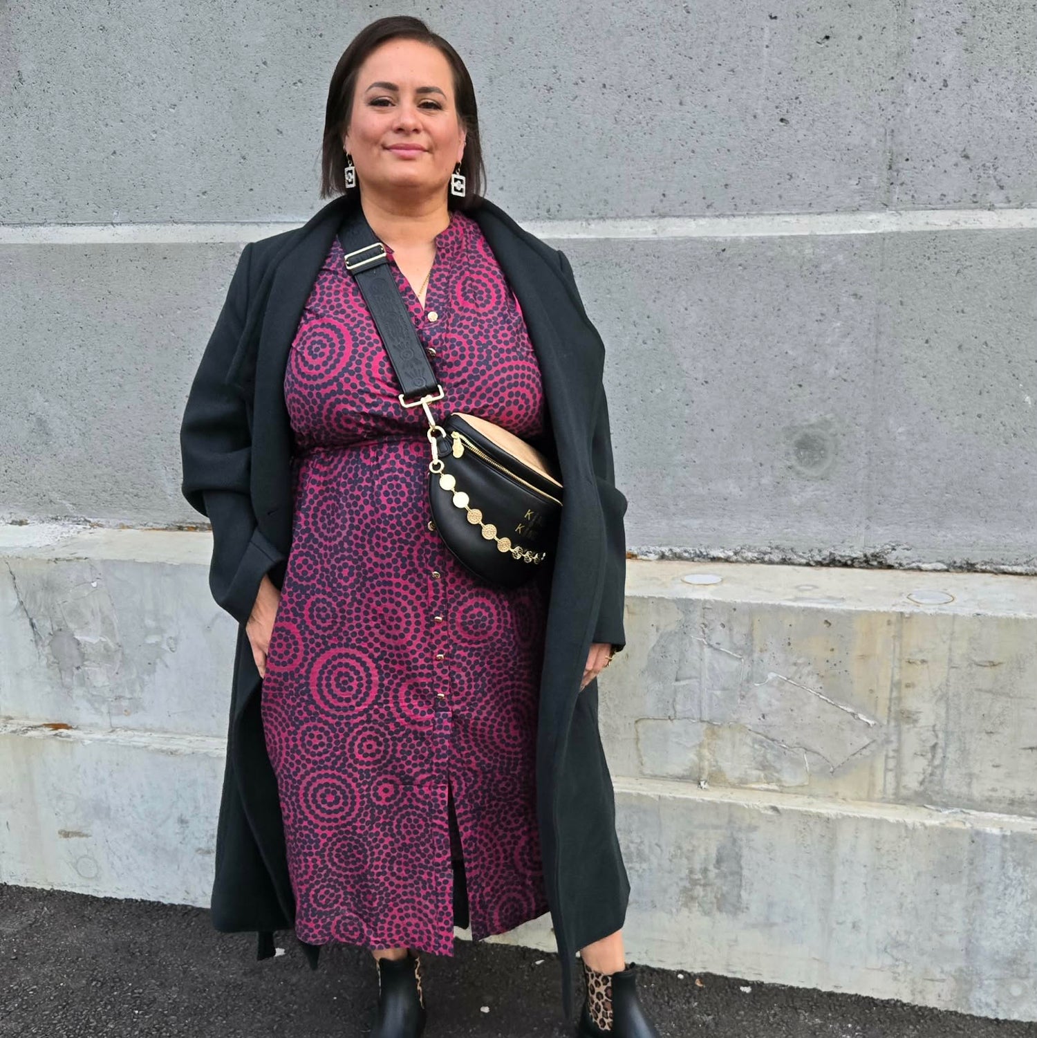 Woman in a patterned dress and long coat standing on steps against a gray wall.